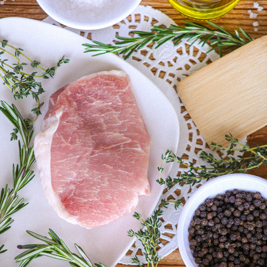 Raw pork chop on a white plate with herbs and peppercorns on a wooden surface