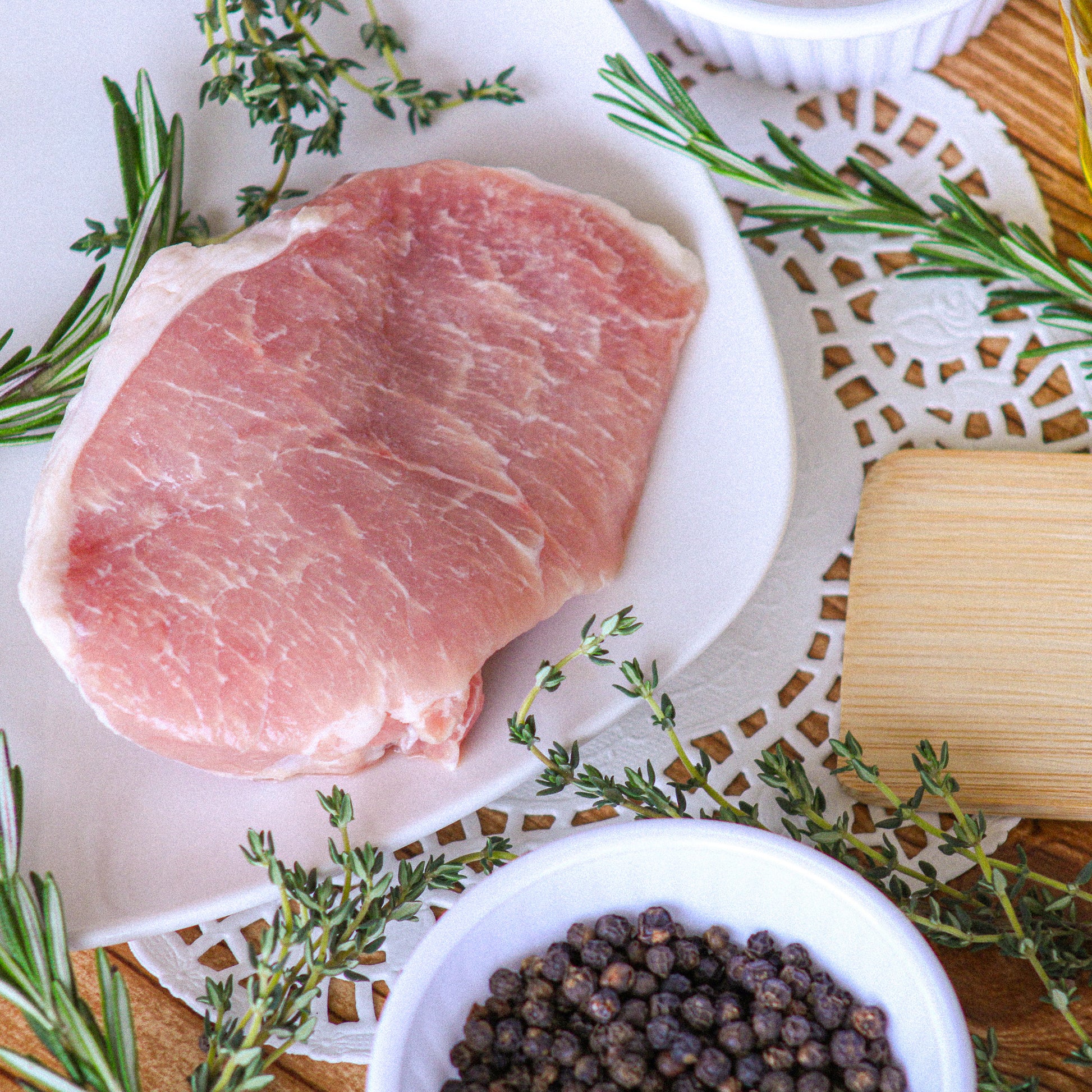 Raw pork chop on a white plate with herbs and black peppercorns on a wooden surface