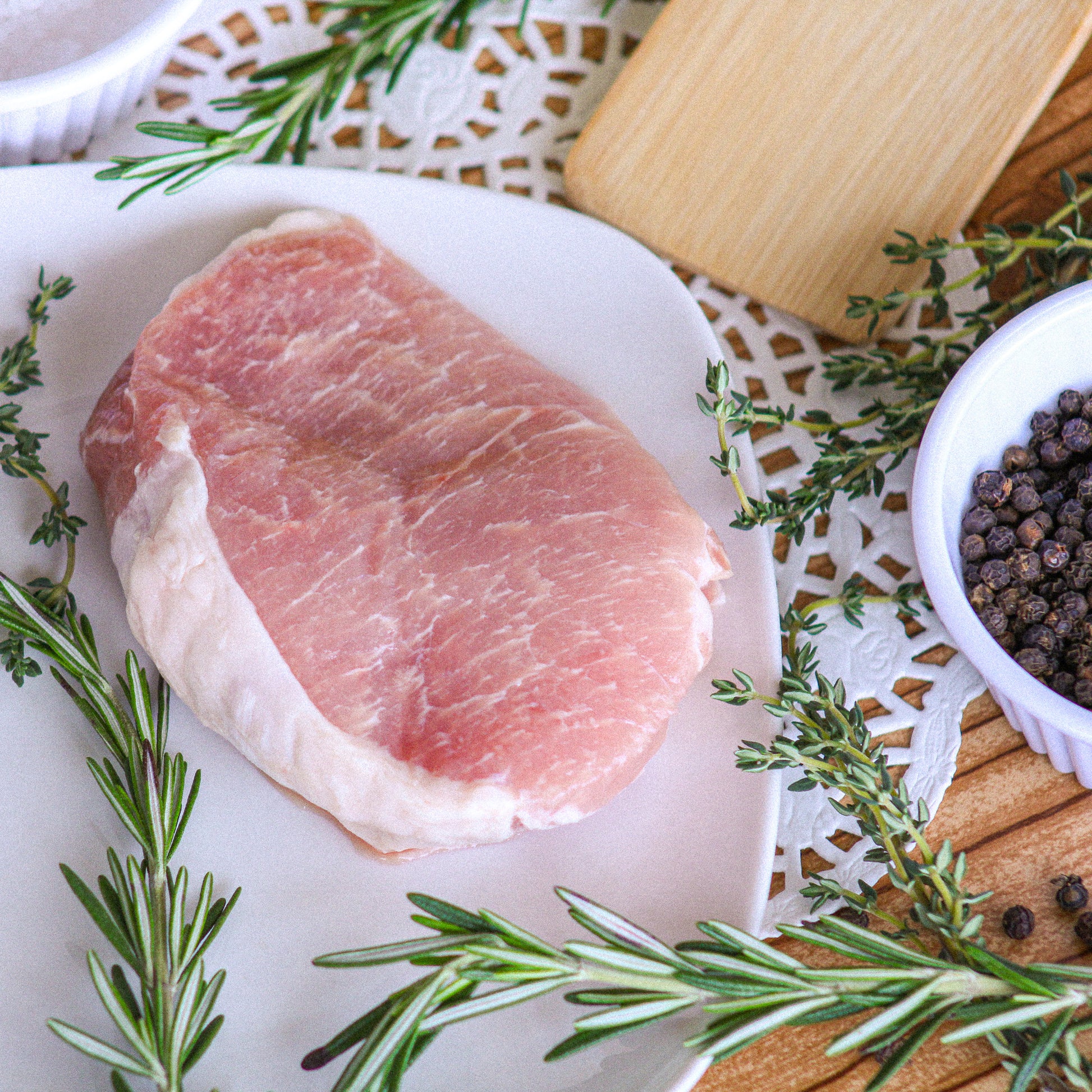 Raw pork chop on a white plate with rosemary and peppercorns.